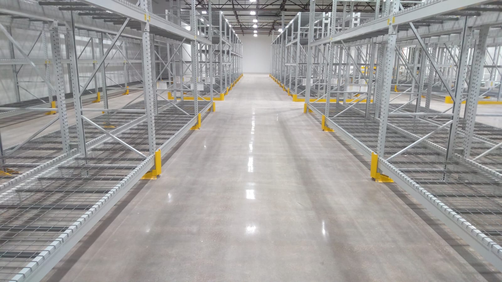 Identical racking bays with wire decking and yellow column protection repeating down both sides of a warehouse aisle
