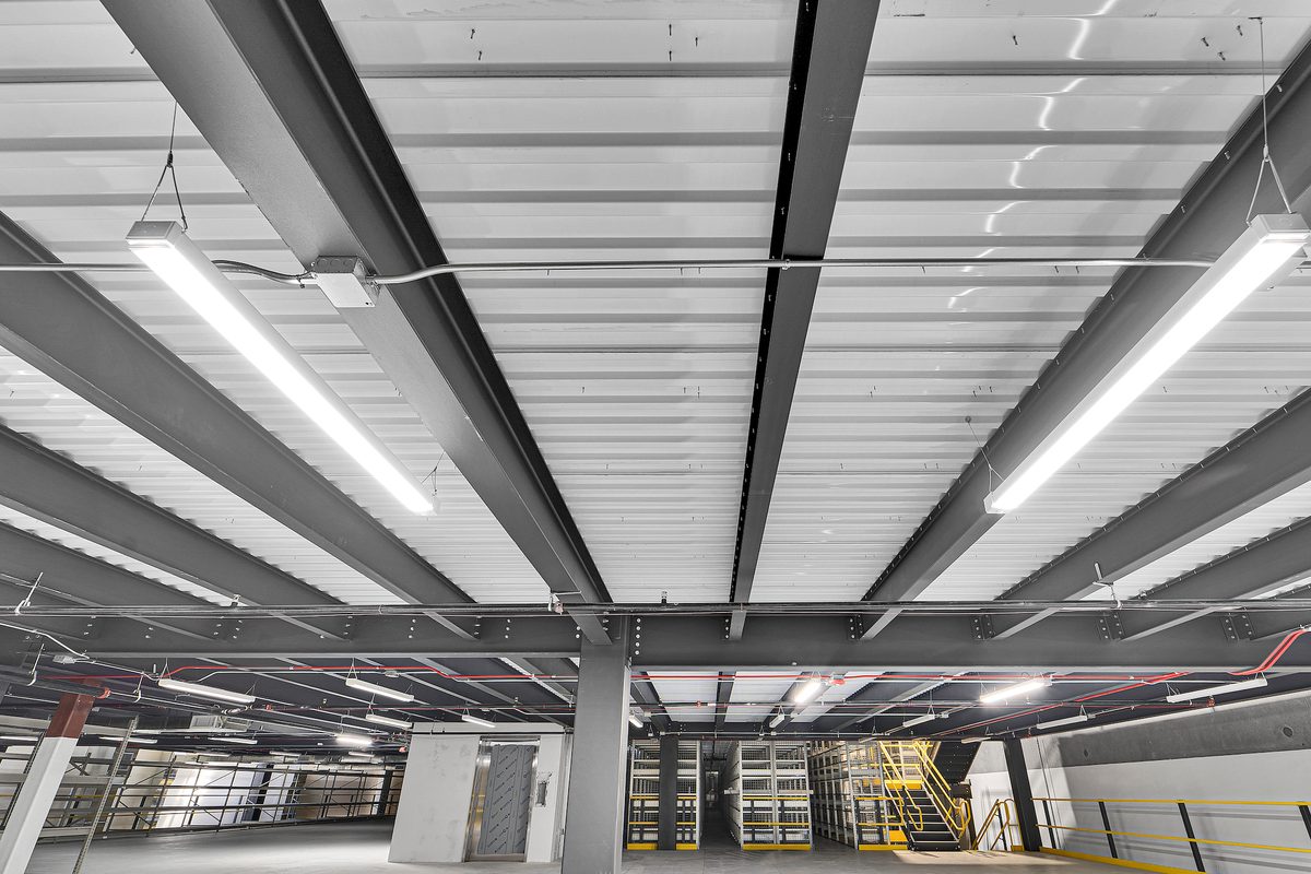 Underside of a structural steel mezzanine: beams and joists carrying a metal deck, with a column bringing the load down to the floor slab